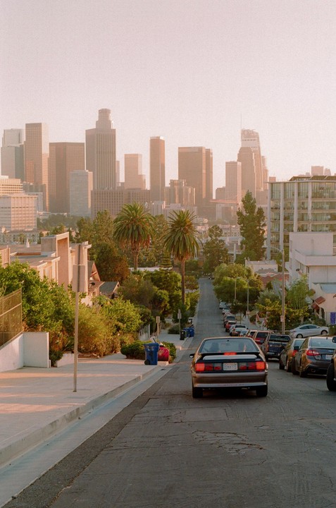A car is parked on a residential street with palm trees and a city skyline in the background during golden hour.