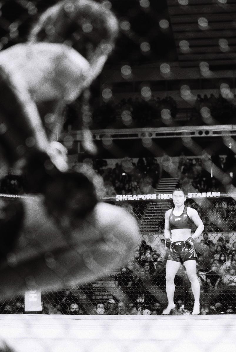 A woman stands in a fighting ring while another fighter is blurred in the foreground.