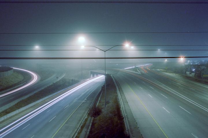 A foggy highway at night shows light trails from vehicles and streetlights illuminating the mist.