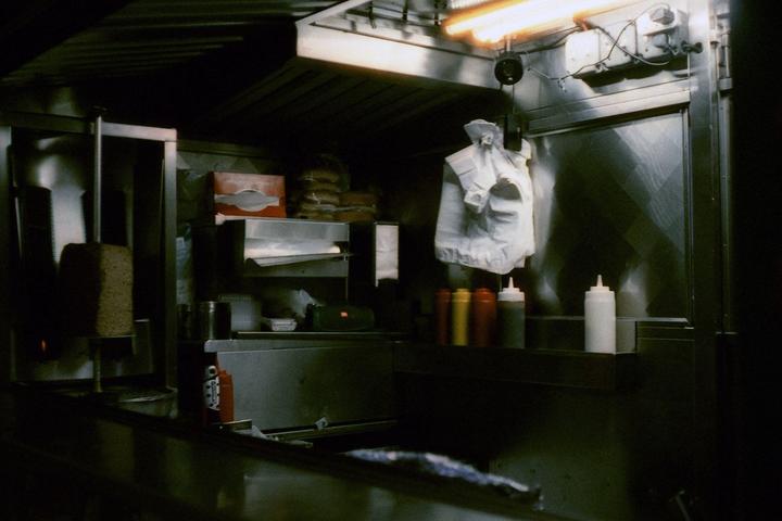 A food truck interior is illuminated by a bright orange light, revealing a vertical rotisserie, condiment bottles, and a hanging white bag.