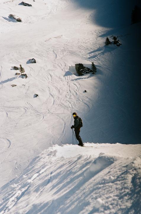 A skier stands on a snowy slope with ski tracks, rocks, and small trees visible in the background.
