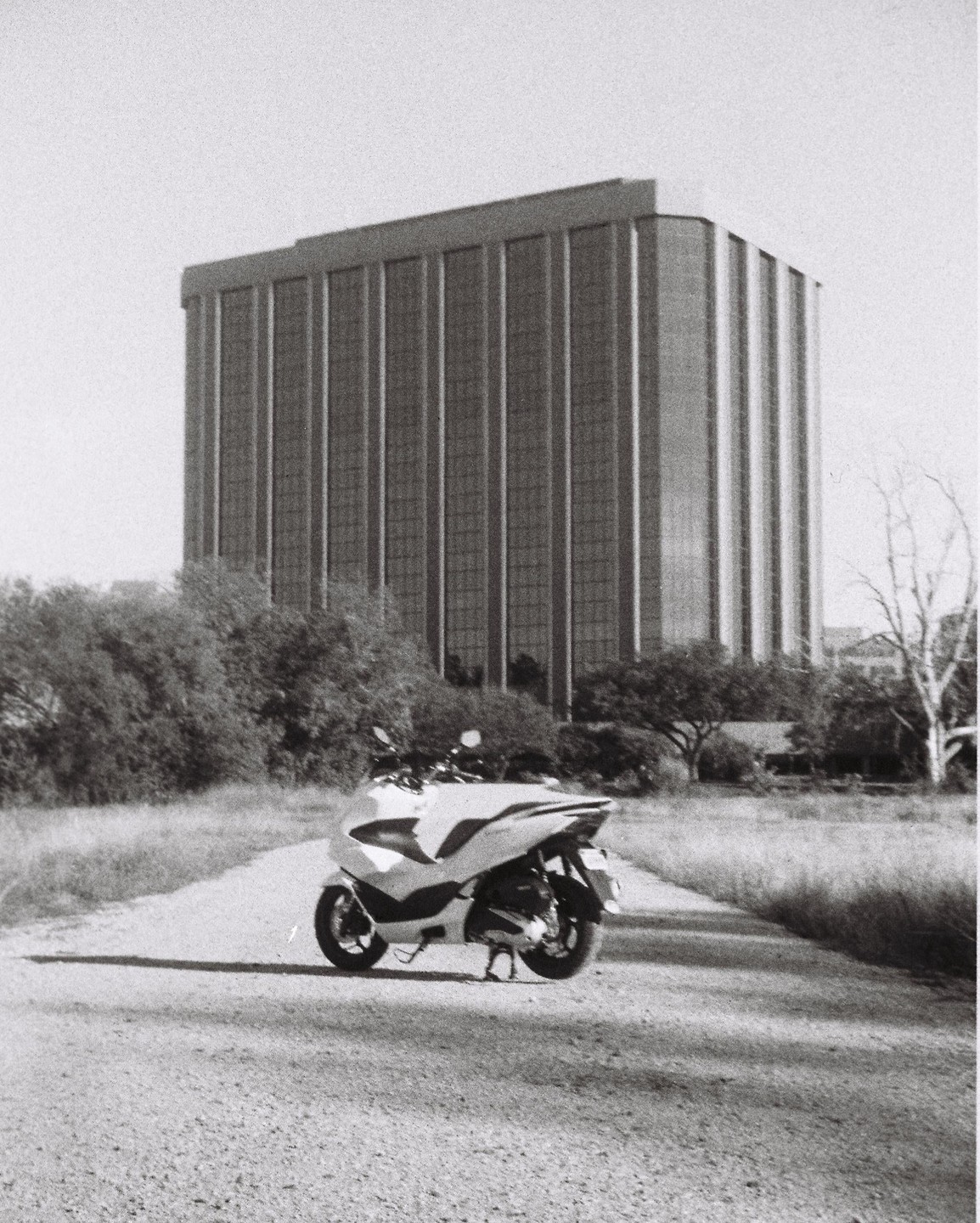 A scooter is parked on a gravel path with a tall building and trees in the background.