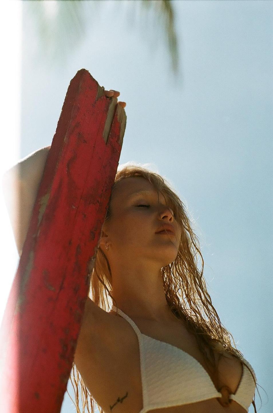 A woman with wet hair and closed eyes leans against a red wooden beam under a bright sky.