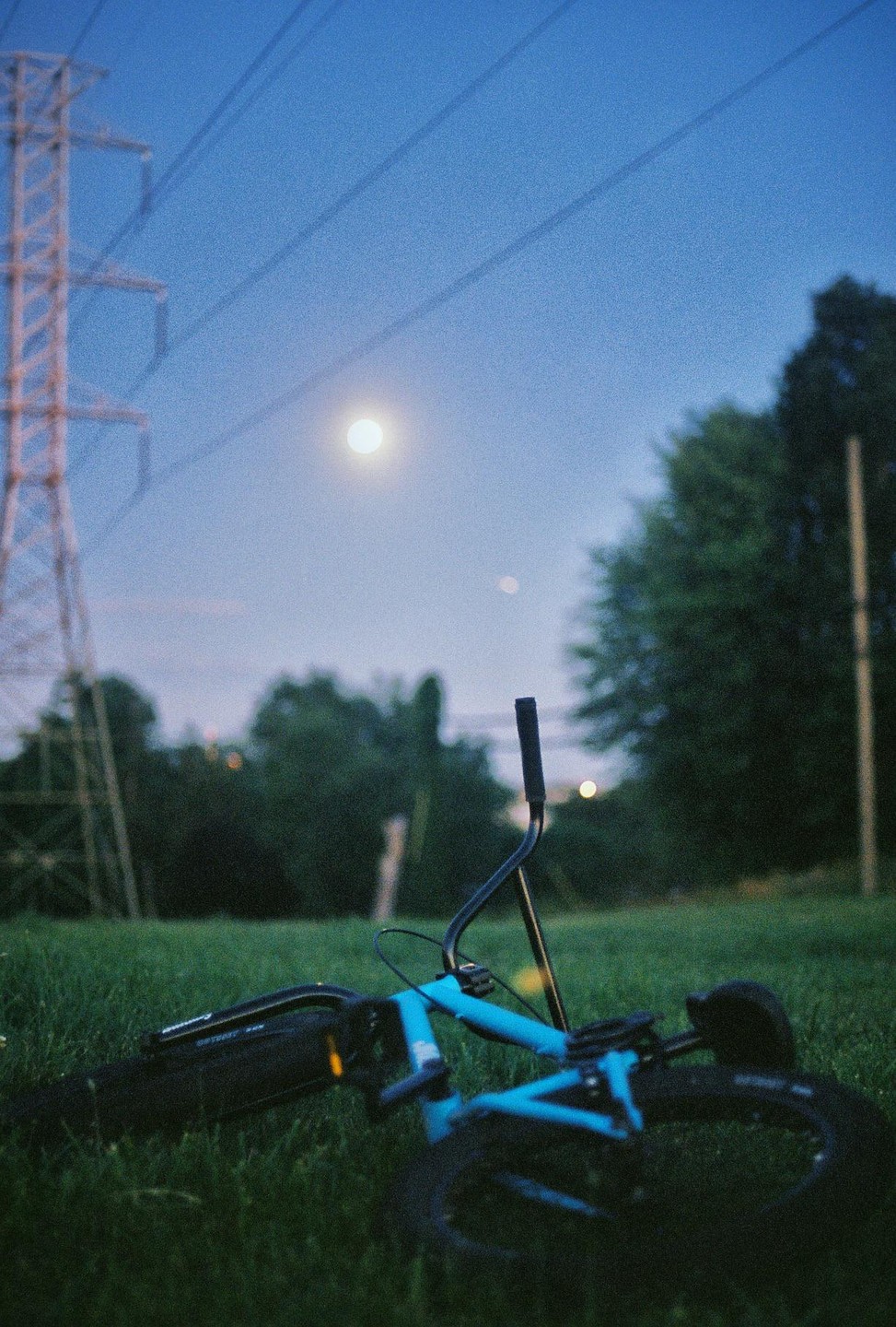 A blue BMX bicycle lies on green grass under a full moon and power lines at dusk.