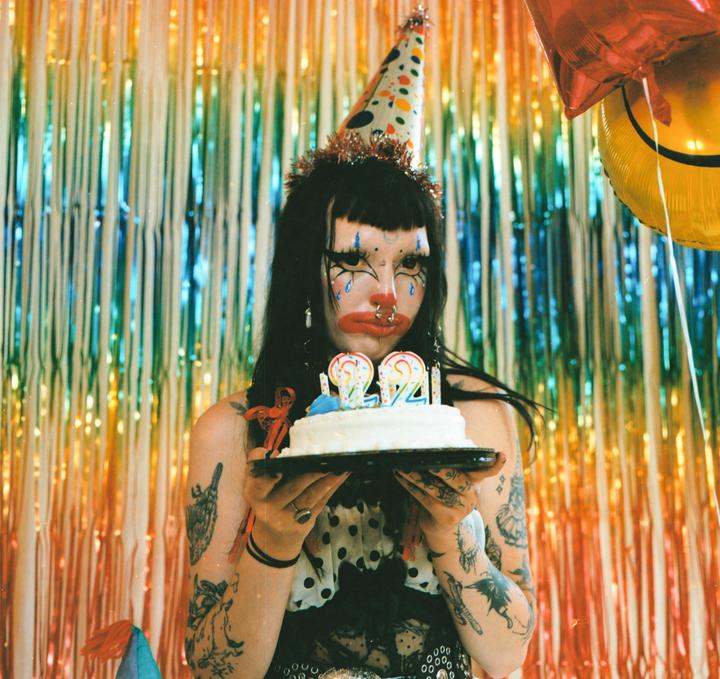 A person with clown makeup and a party hat holds a birthday cake with candles in front of a rainbow tinsel curtain.