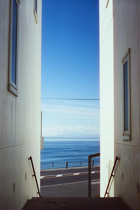 A narrow view between two buildings reveals a road, the ocean, and a distant mountain range under a clear blue sky.