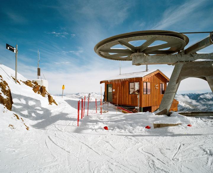 A wooden building with icicles on its roof stands next to a large ski lift mechanism on a snow-covered mountain under a blue sky.