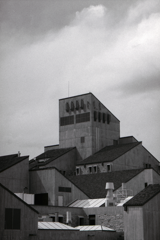 A tall concrete building with a slanted roof stands among other buildings with various rooflines under a cloudy sky.