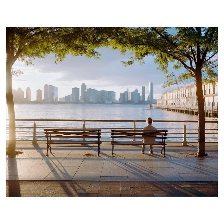 A person sits on a park bench looking out at a river with a city skyline in the distance.