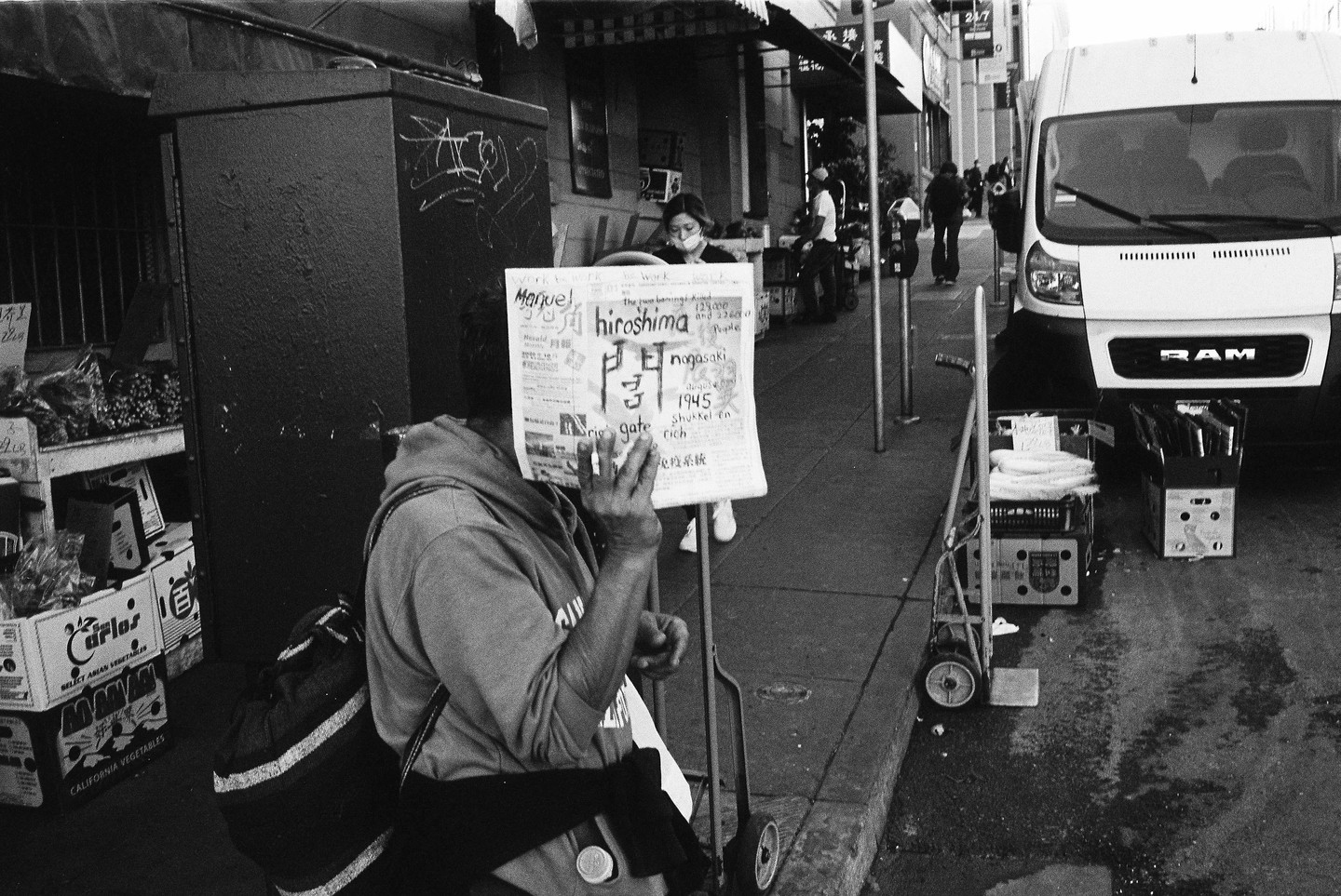 A person holds a newspaper with the headline "Hiroshima" on a city street next to a white van.