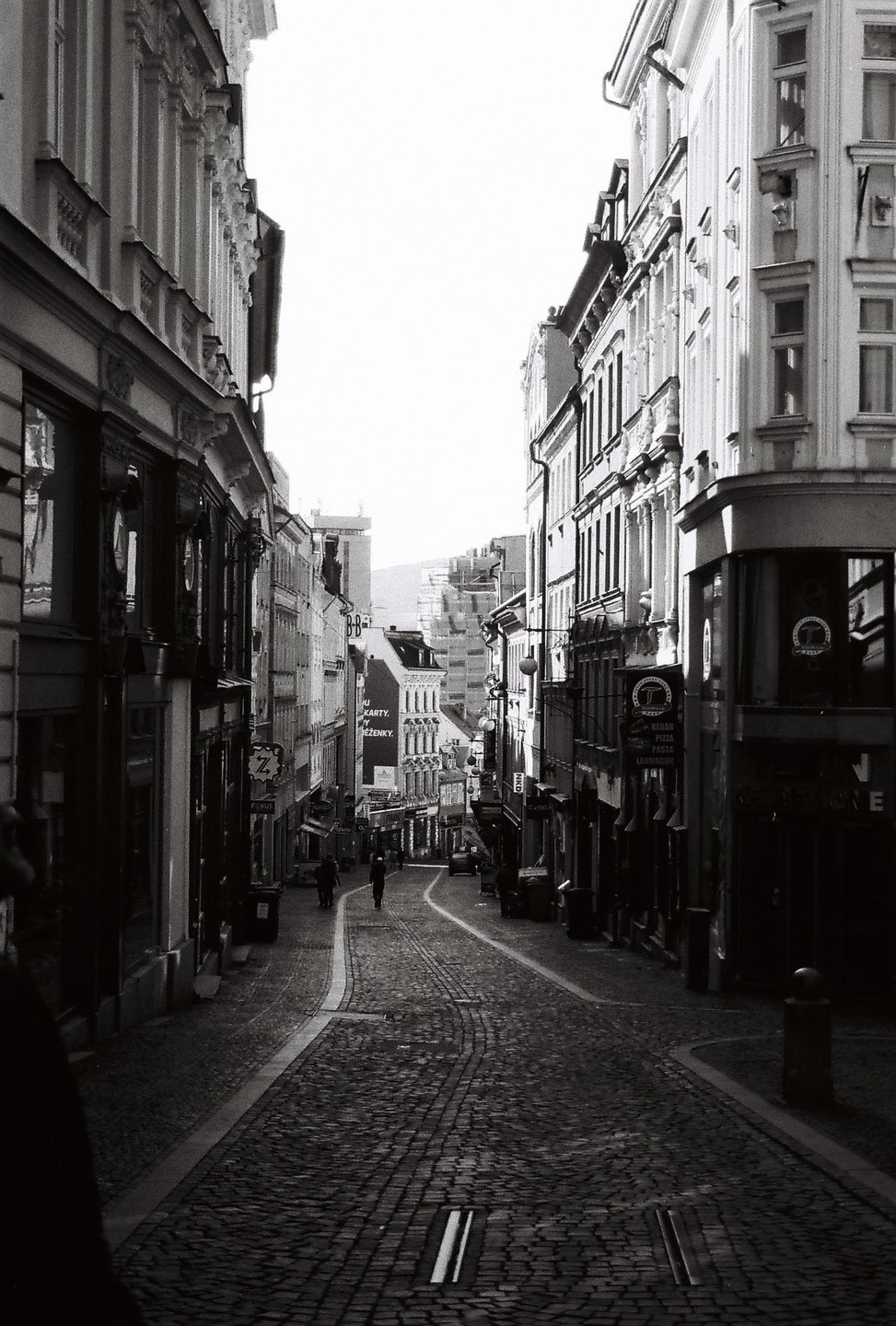 A narrow, cobbled street lined with tall buildings leads into the distance, with a few people walking along it.