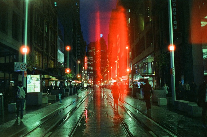 A street at night is wet from rain, with tram tracks running down the middle and red light flares across the image.