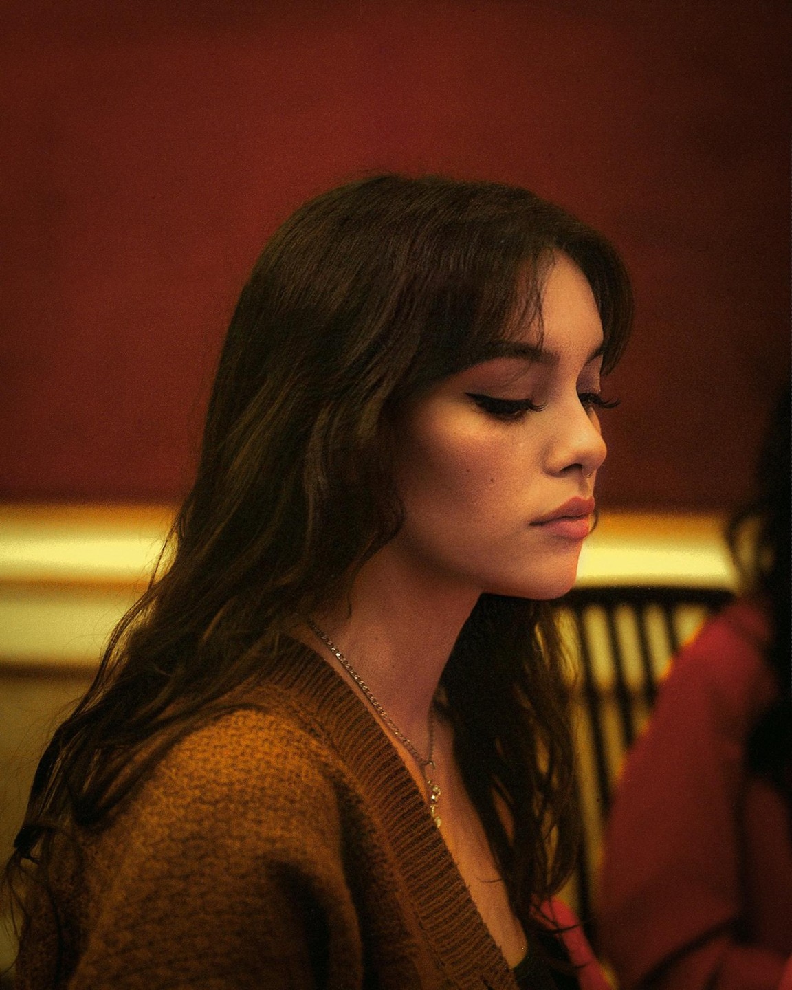 A woman with long dark hair and a brown cardigan looks down, illuminated by warm light.