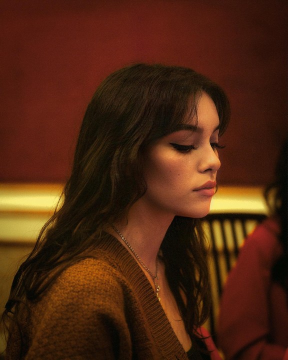 A woman with long dark hair and a brown cardigan looks down, illuminated by warm light.