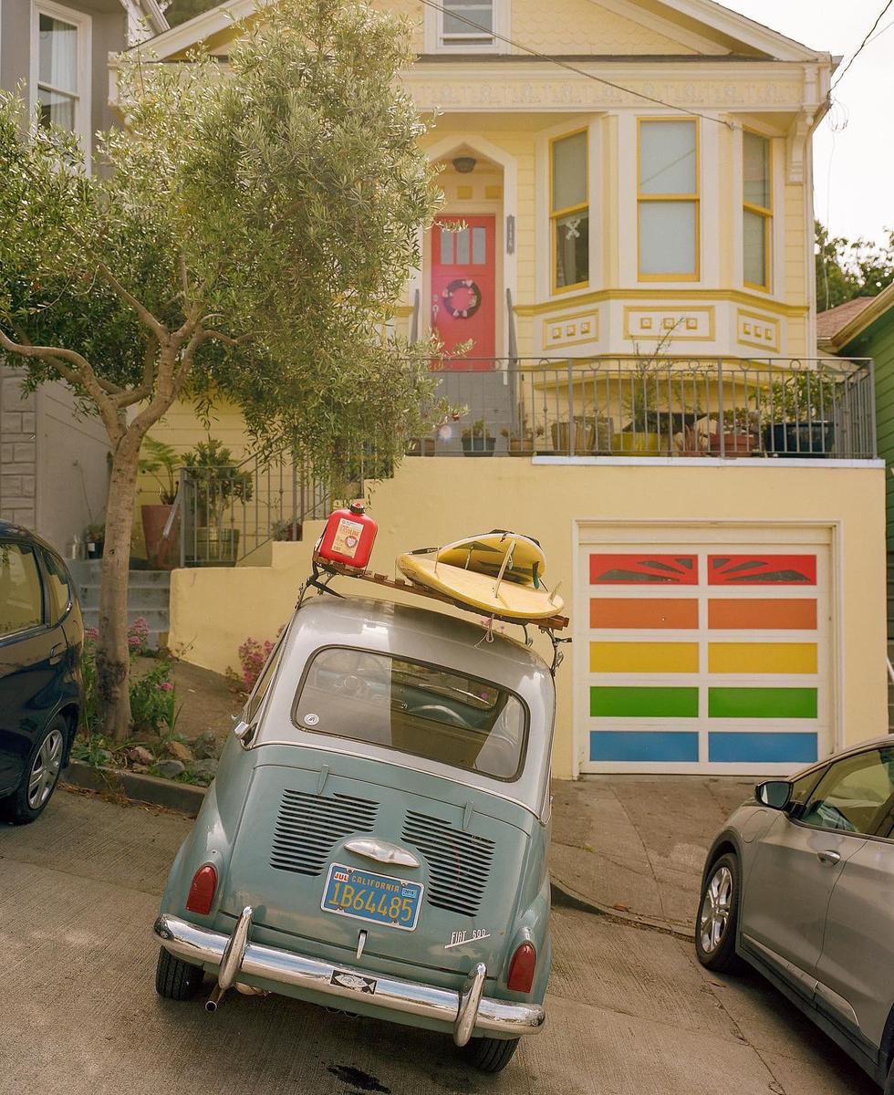 A small car with a surfboard and gas can on its roof is parked on a steep street in front of a yellow house with a rainbow-colored garage door.