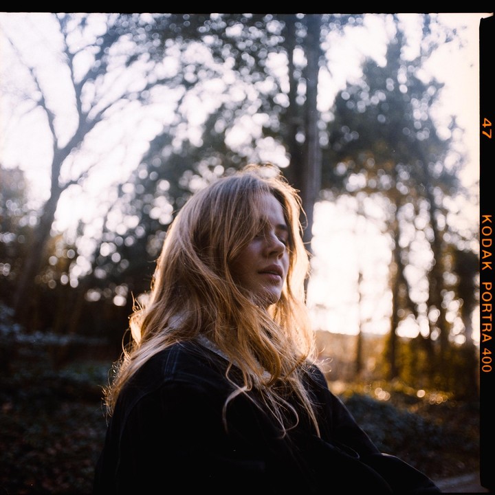 A woman with long hair and closed eyes is backlit by the setting sun in a forest.