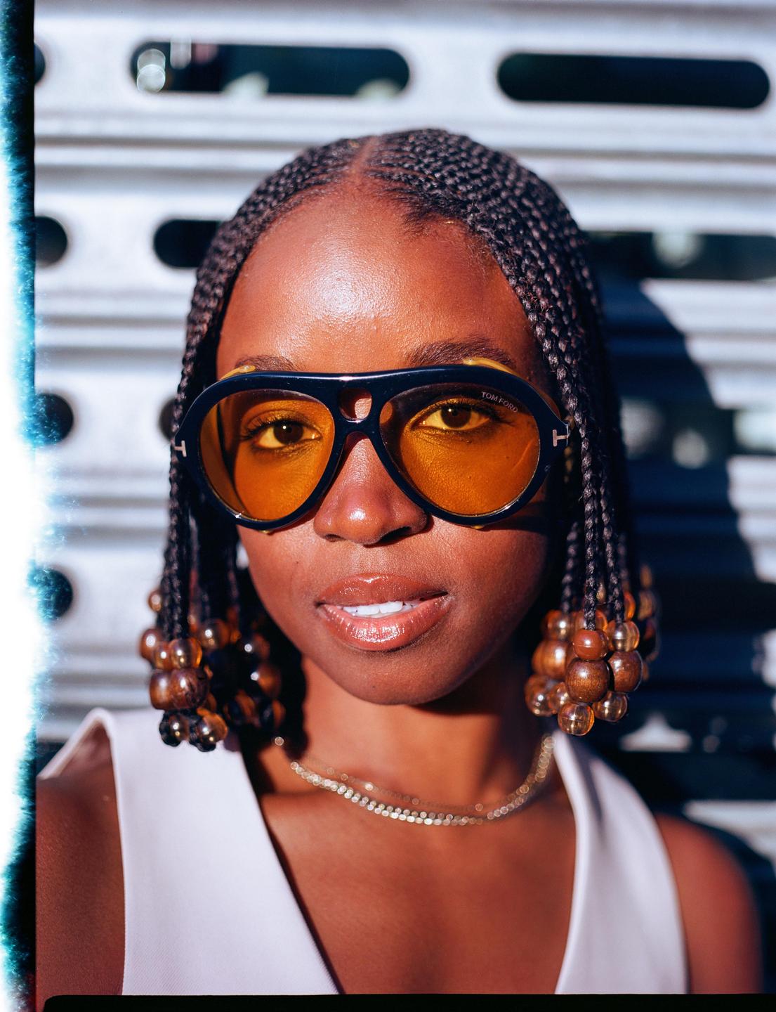 A woman with braided hair and sunglasses looks at the viewer.