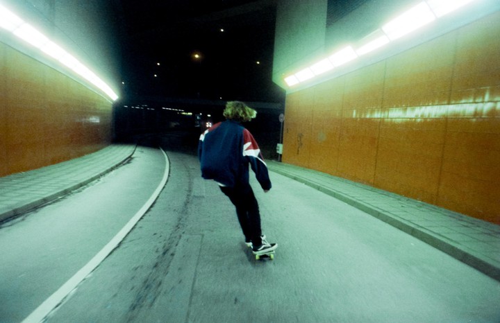 A person with curly hair skates away from the viewer in a tunnel at night.