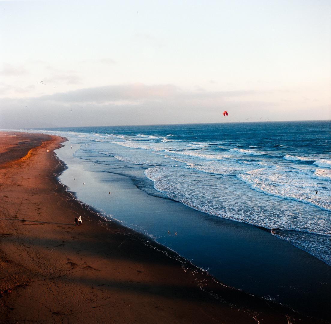 A red kite flies over the ocean next to a sandy beach with waves crashing on the shore.