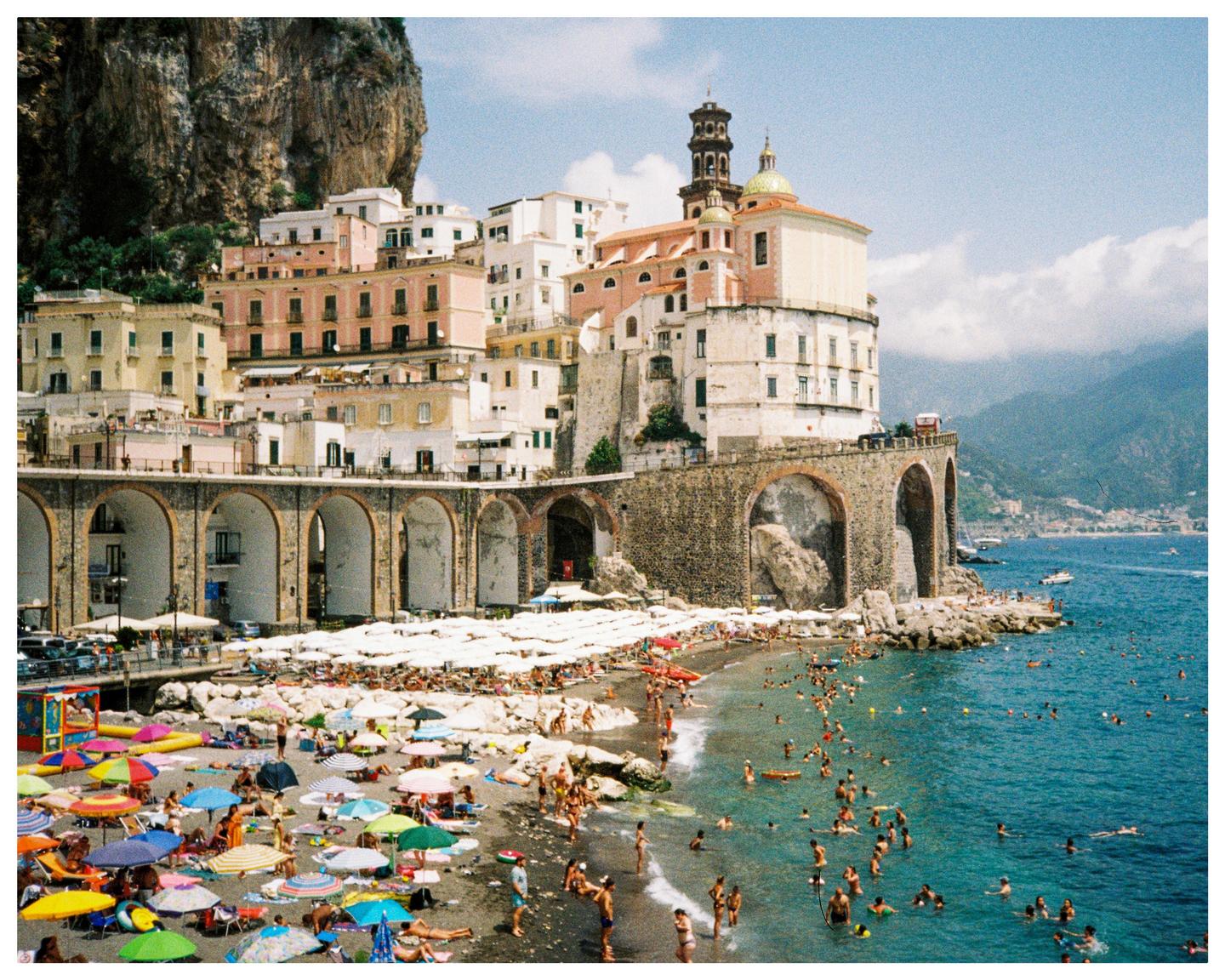 A crowded beach with many colorful umbrellas is next to a town built into a cliffside overlooking the sea.