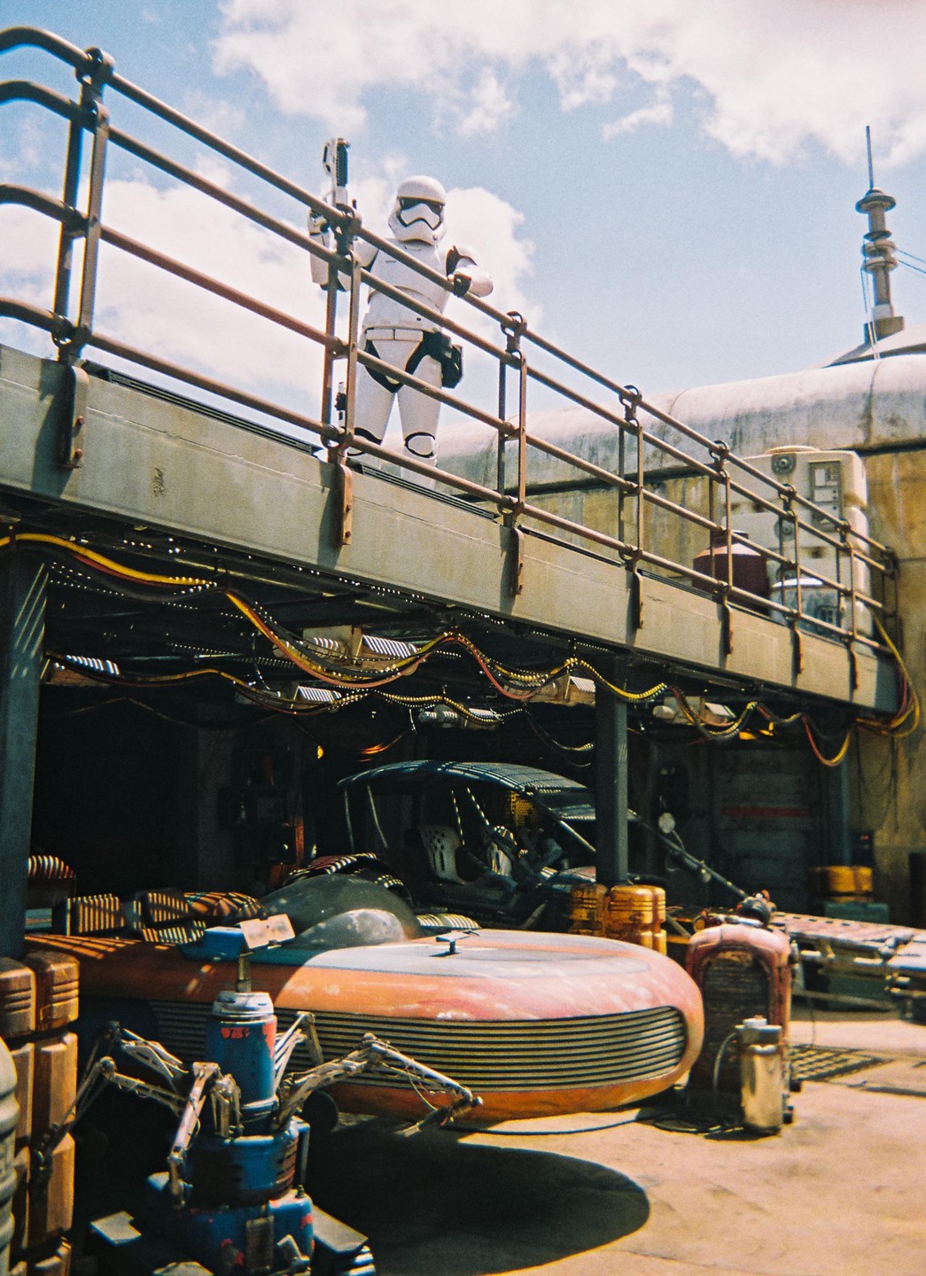 A stormtrooper stands on a metal walkway above a futuristic vehicle and other structures under a cloudy sky.