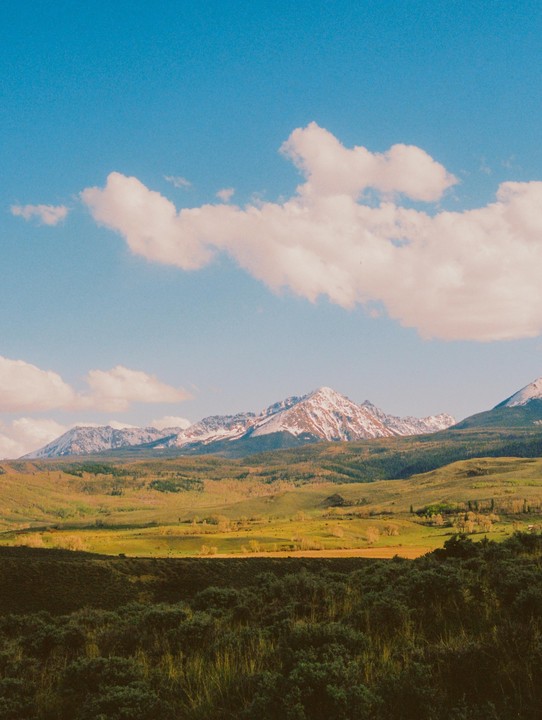 A vast landscape features snow-capped mountains in the distance, rolling green hills, and a field of dark green shrubs in the foreground under a blue sky with white clouds.