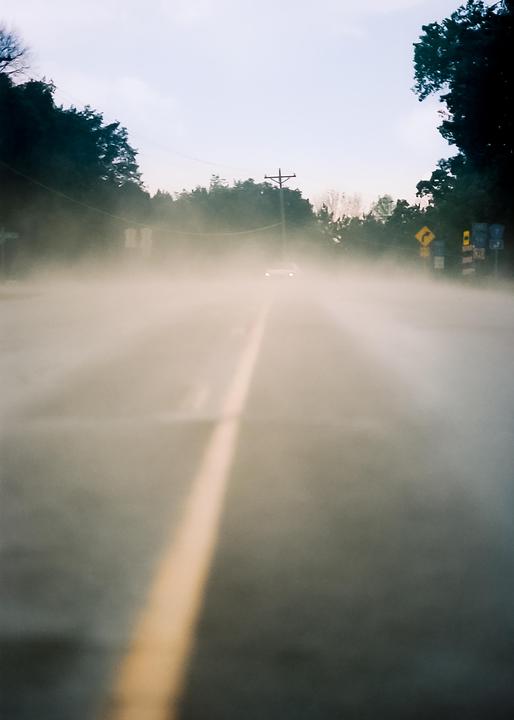 A car with its headlights on drives down a hazy road with a yellow dividing line.