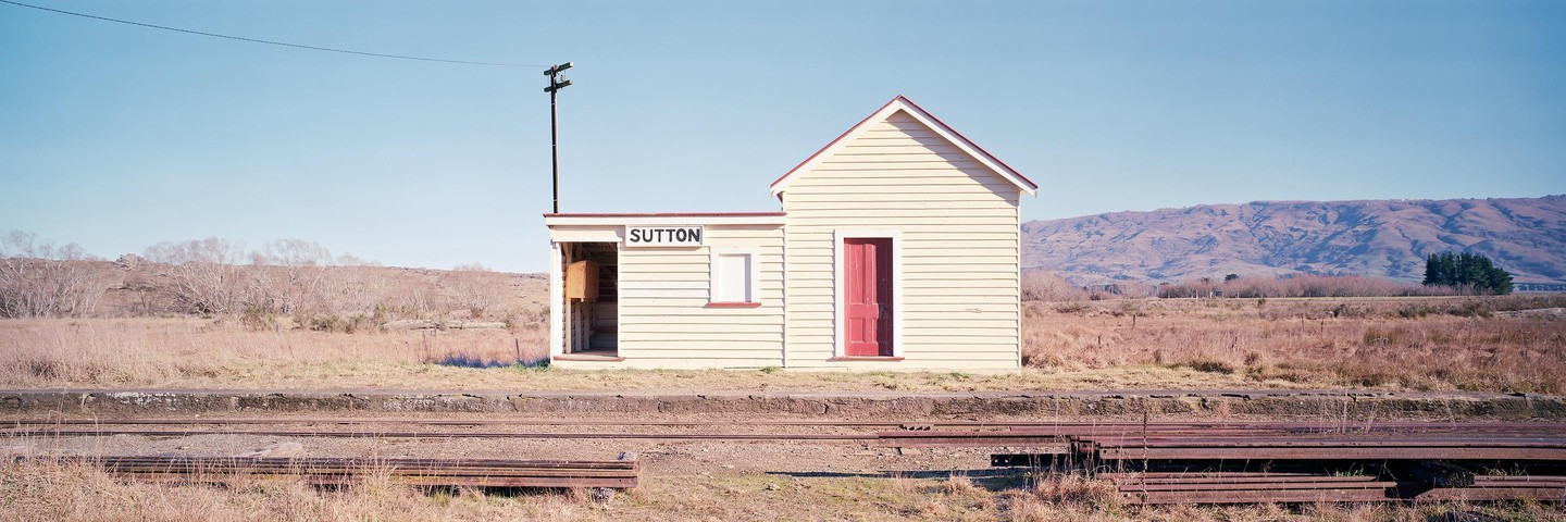 A small, light-colored building with a red door and a sign that says "SUTTON" stands next to train tracks in a dry, grassy landscape with mountains in the background.