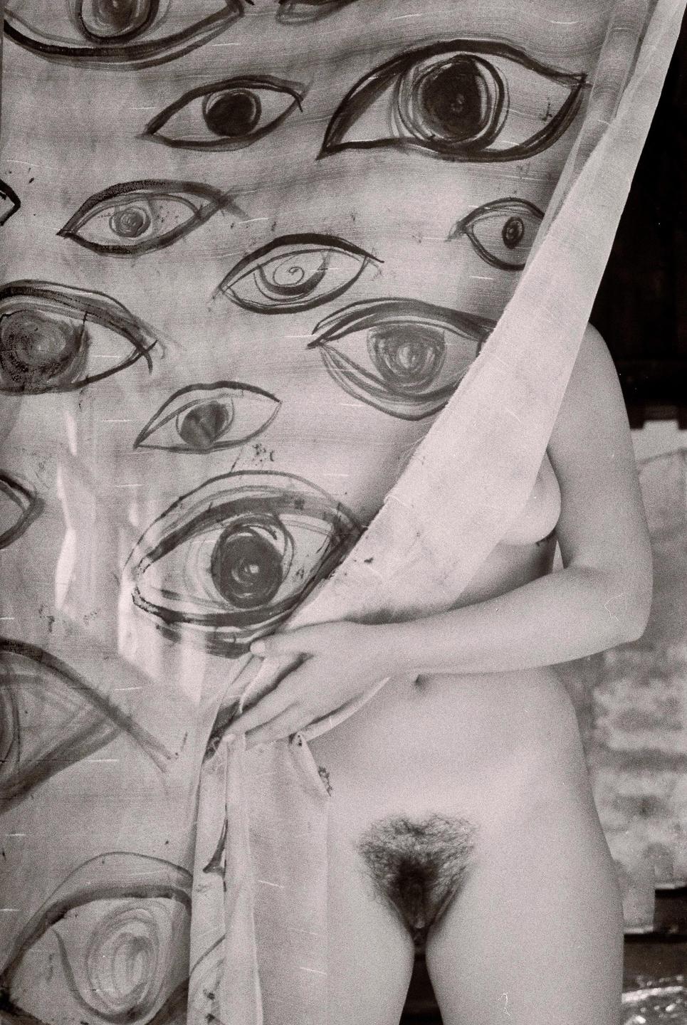 A woman stands partially nude behind a sheer fabric with painted eyes.