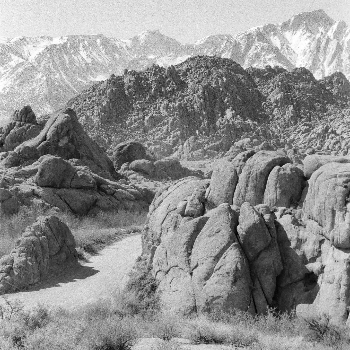 A dirt road winds through a rocky landscape with snow-capped mountains in the background.