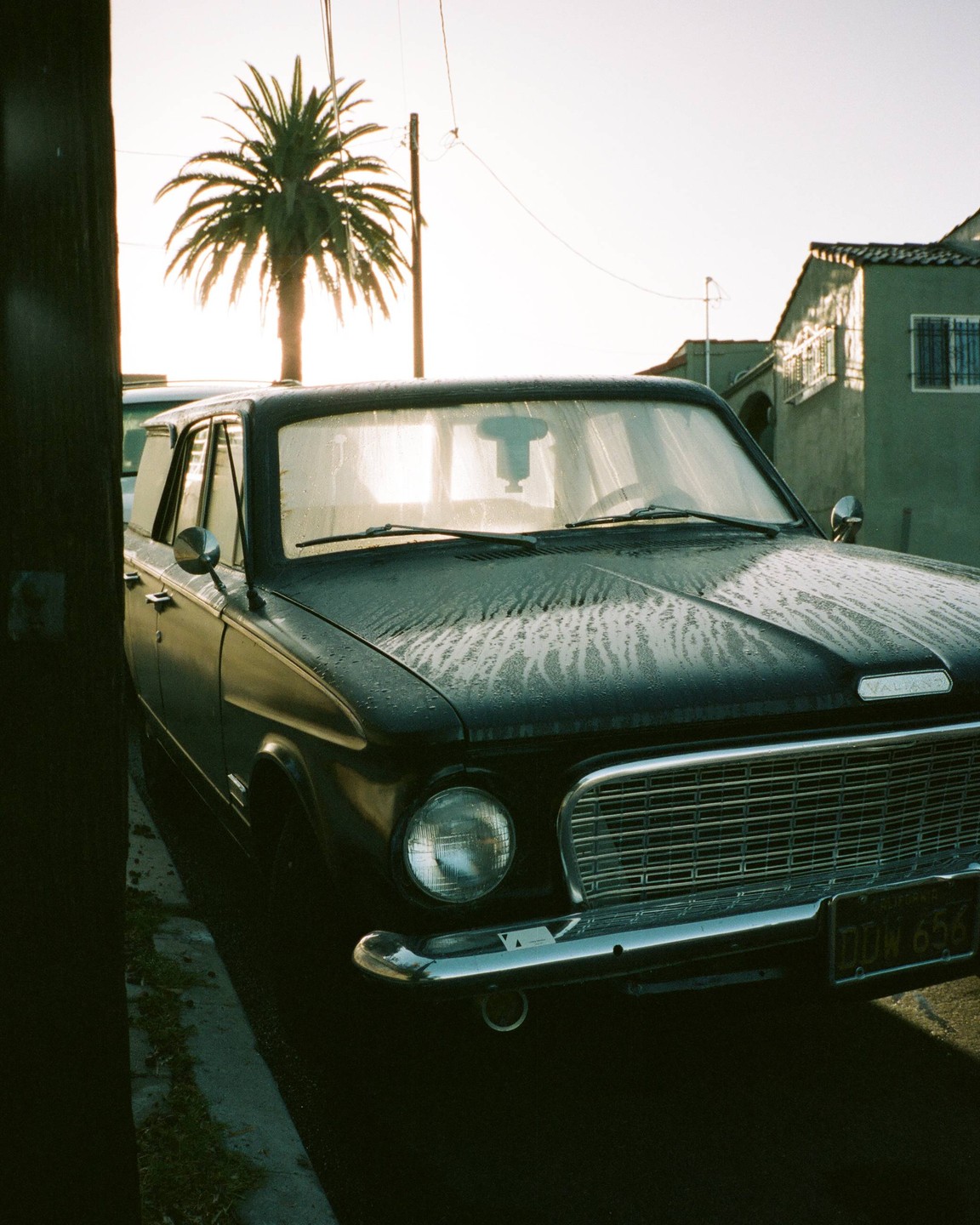 A black vintage car with water streaks on its hood is parked on a street with a palm tree and building in the background.