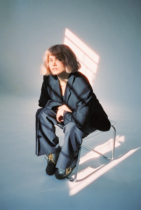A woman with short hair and a dark suit sits on a clear chair with window light and shadow falling on her and the floor.