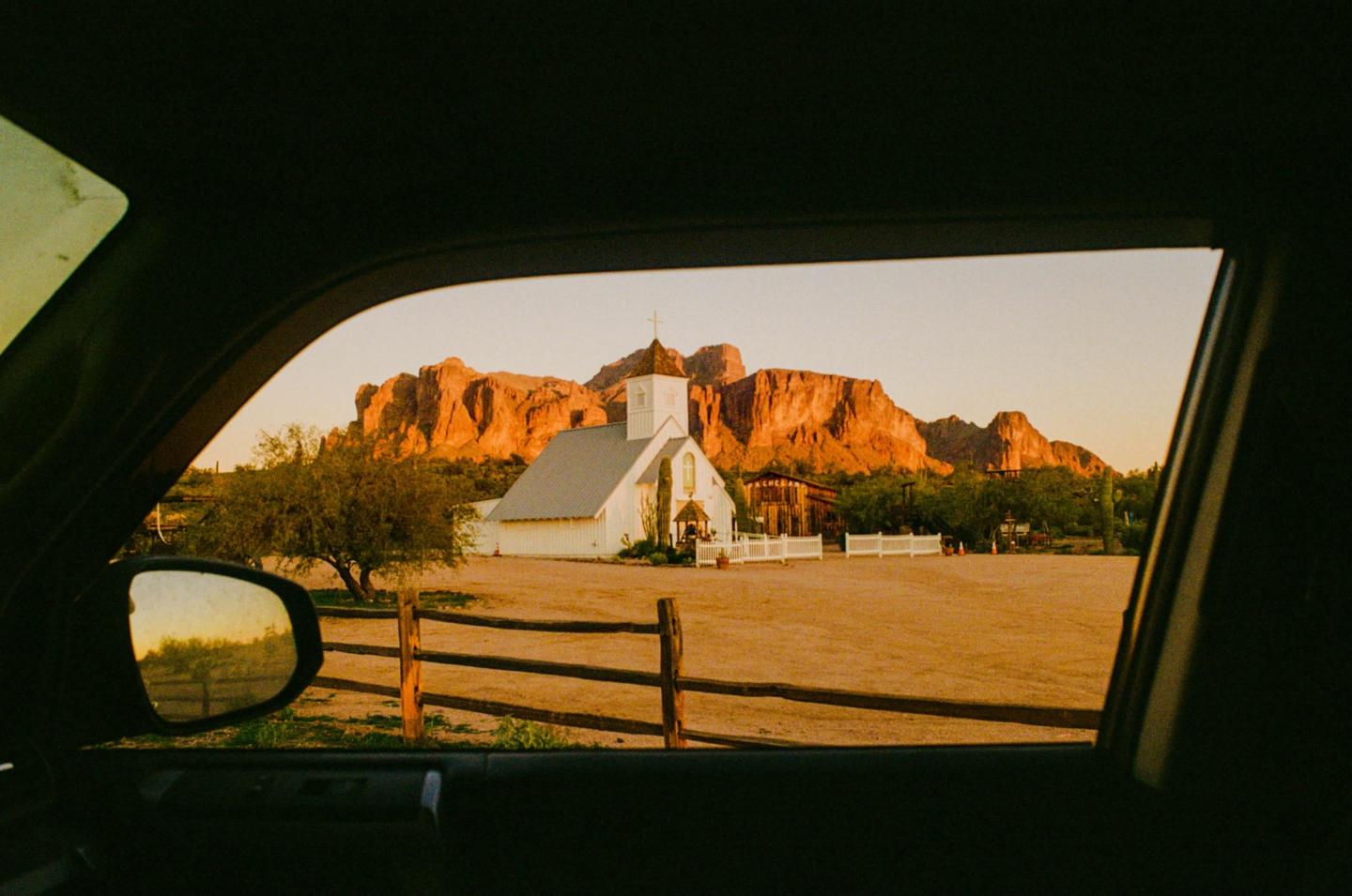 A white church with a steeple stands in a desert landscape with mountains in the background, viewed through a car window.