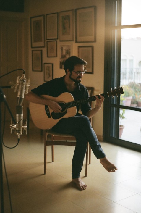 A man sits on a chair playing an acoustic guitar next to a microphone and a window.