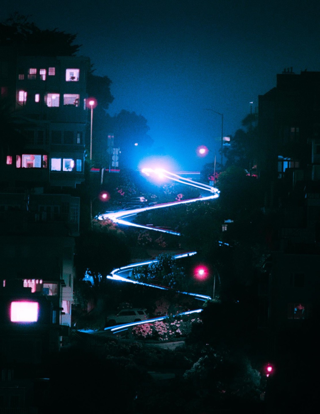 A long exposure shot captures the light trails of vehicles on a winding street at night, with buildings and trees visible on either side.