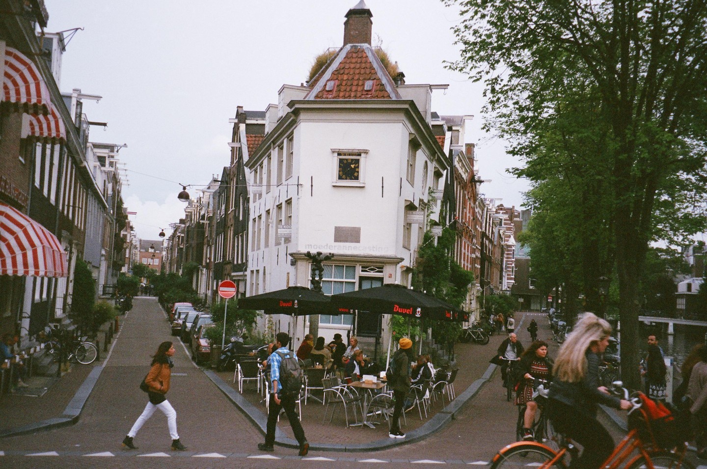 People walk and cycle on a street with buildings and trees, with a cafe on the corner.