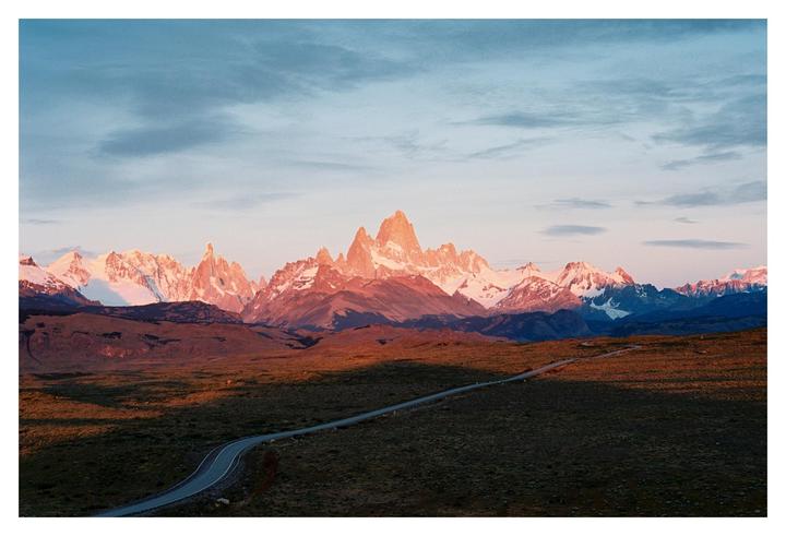 A winding road leads towards a mountain range with snow-capped peaks illuminated by the setting sun.