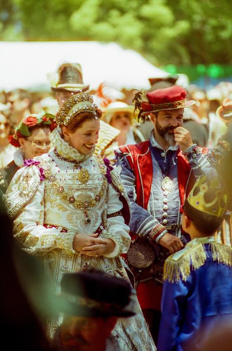 A woman in elaborate period costume and a man in a red hat and period costume look at a child in a blue costume and gold crown.