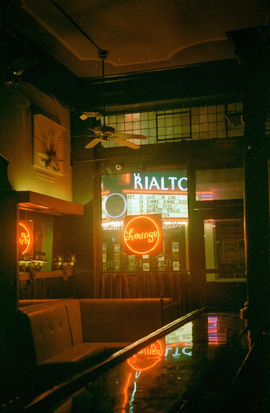 A neon "Lounge" sign is reflected in a wet surface inside a building with a ceiling fan.