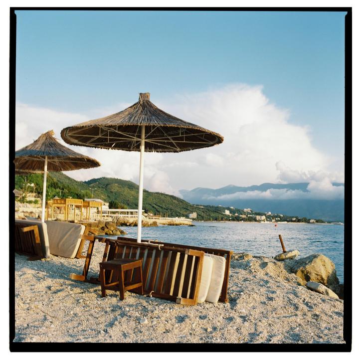Two straw umbrellas stand on a rocky beach next to the sea, with mountains and a town in the background.
