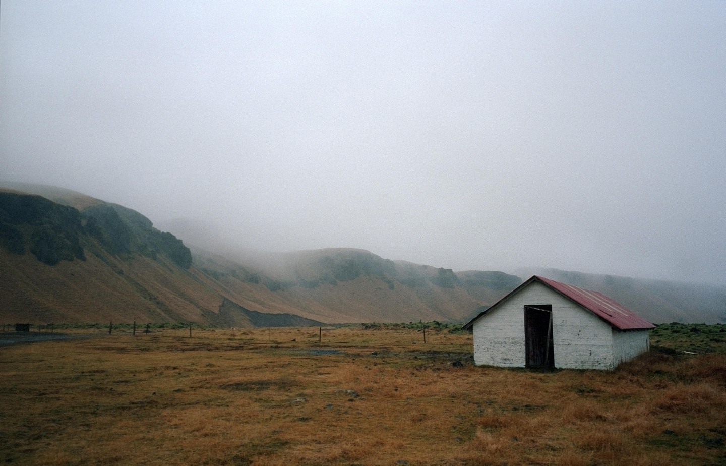 A small white building with a red roof stands in a field with a mountain in the background on a foggy day.