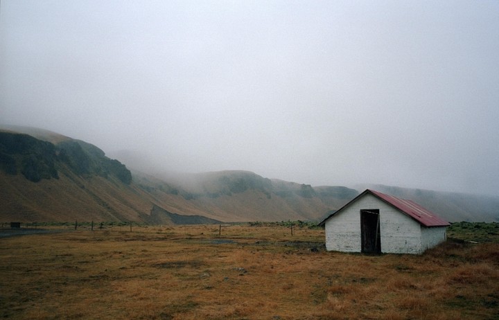 A small white building with a red roof stands in a field with a mountain in the background on a foggy day.