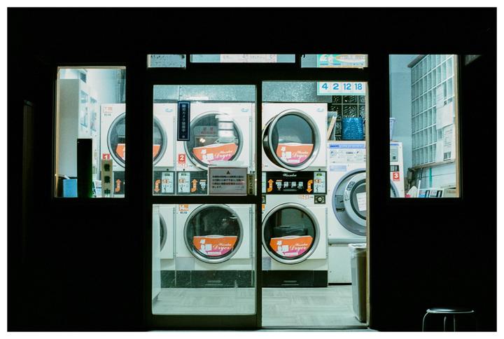A row of washing machines and dryers are visible through the glass doors and windows of a laundromat at night.