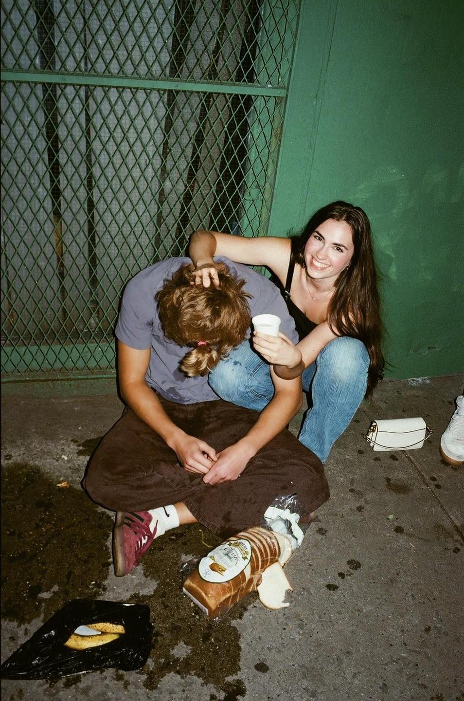 A woman smiles at the camera while a man sits with his head down next to her on a street at night.