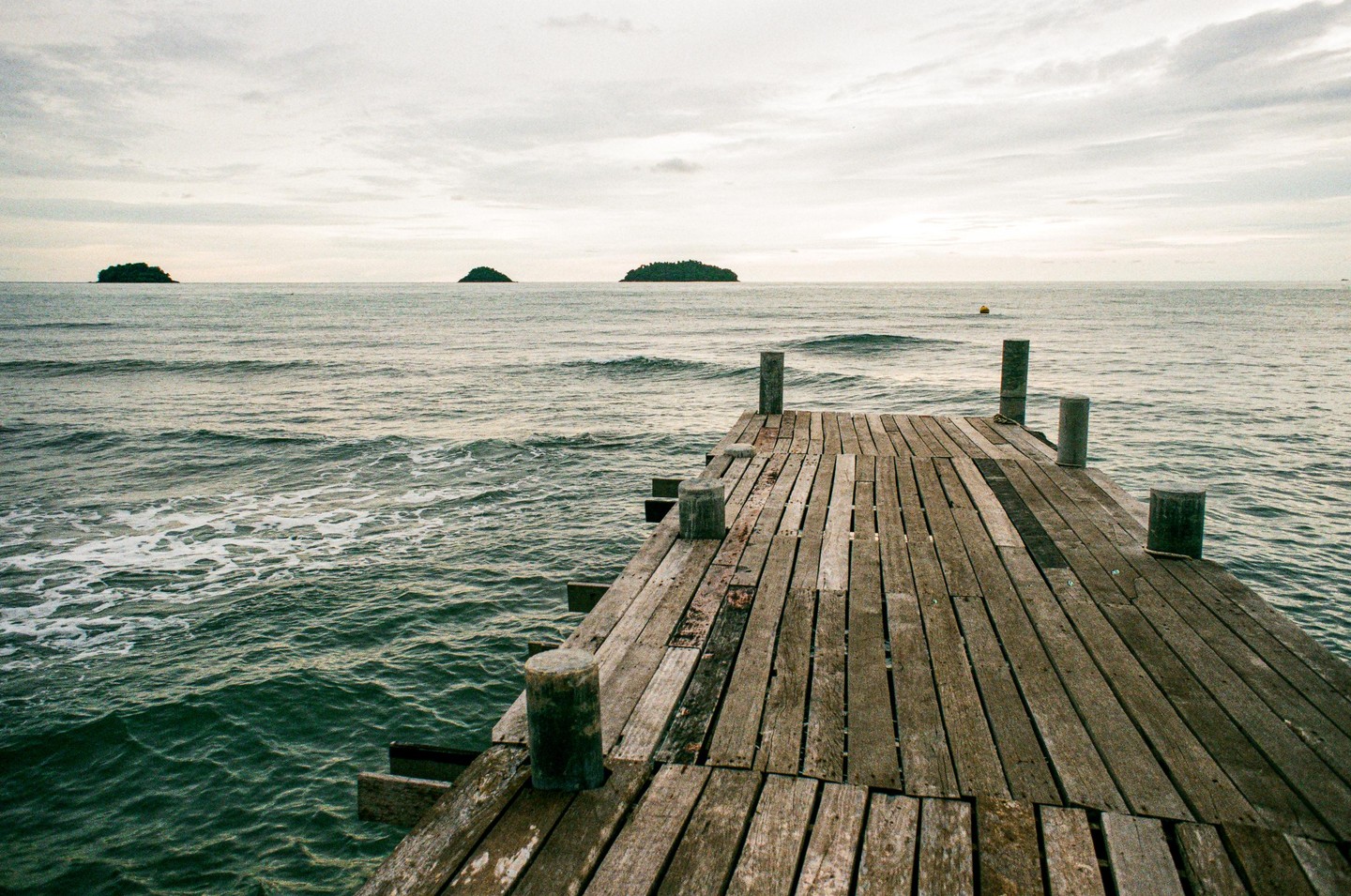 A wooden pier extends into the ocean with small islands visible in the distance under a cloudy sky.