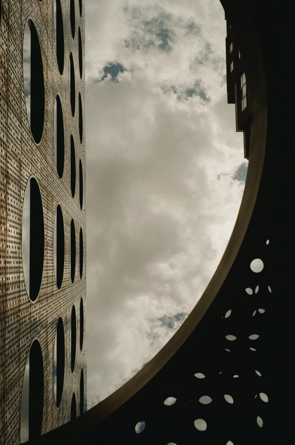 A low-angle view shows a modern building with circular windows and a curved structure with small cutouts against a cloudy sky.