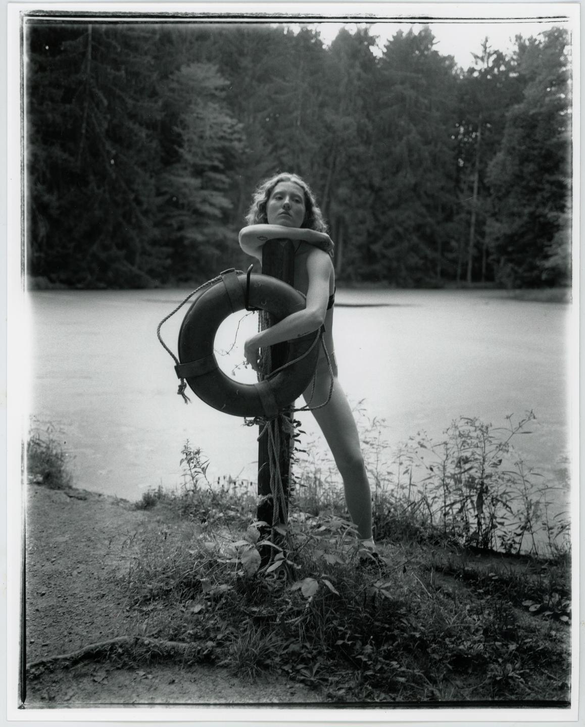 A woman in a swimsuit leans on a wooden post with a life preserver by a lake surrounded by a forest.