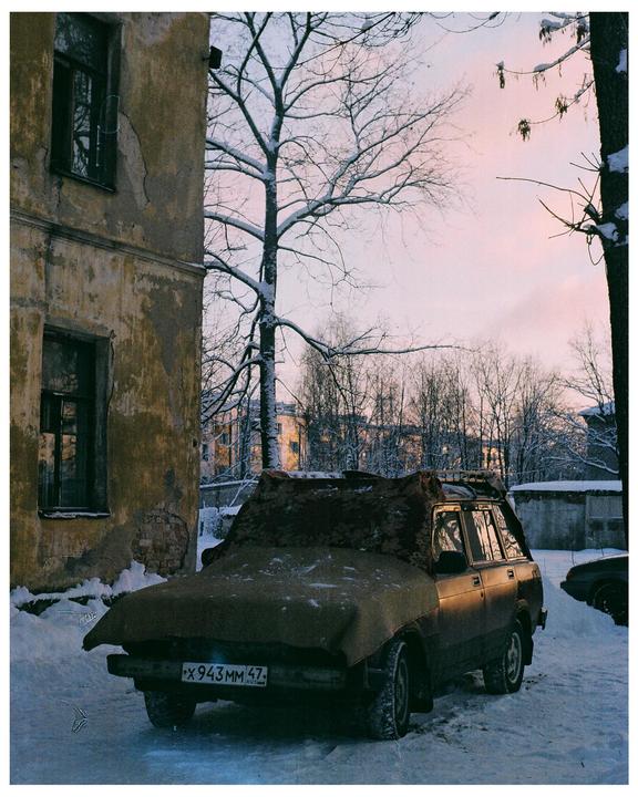 A car covered with a tarp is parked in the snow next to a building with a bare tree in the background.