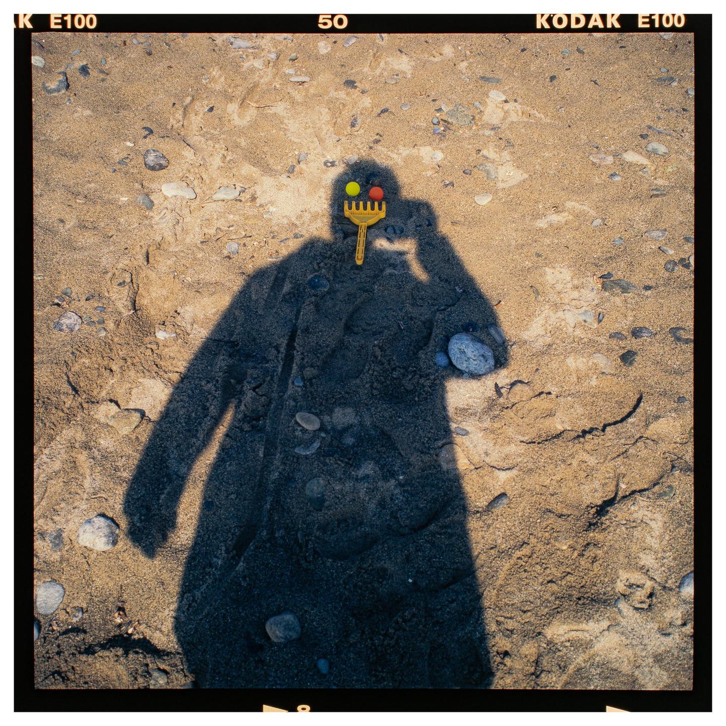A shadow of a person holding a toy rake with colorful balls on it is cast on a sandy beach.