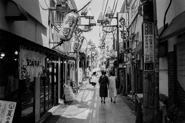 Three women walk away from the viewer down a narrow street lined with shops and signs.